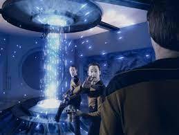 Three versions of Data standing a few feet apart near a vertical energy beam; one has turned toward the viewer and is speaking